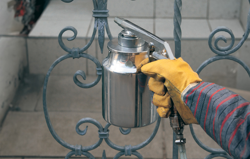 A person is sandblasting a metal railing using a sand spray gun.