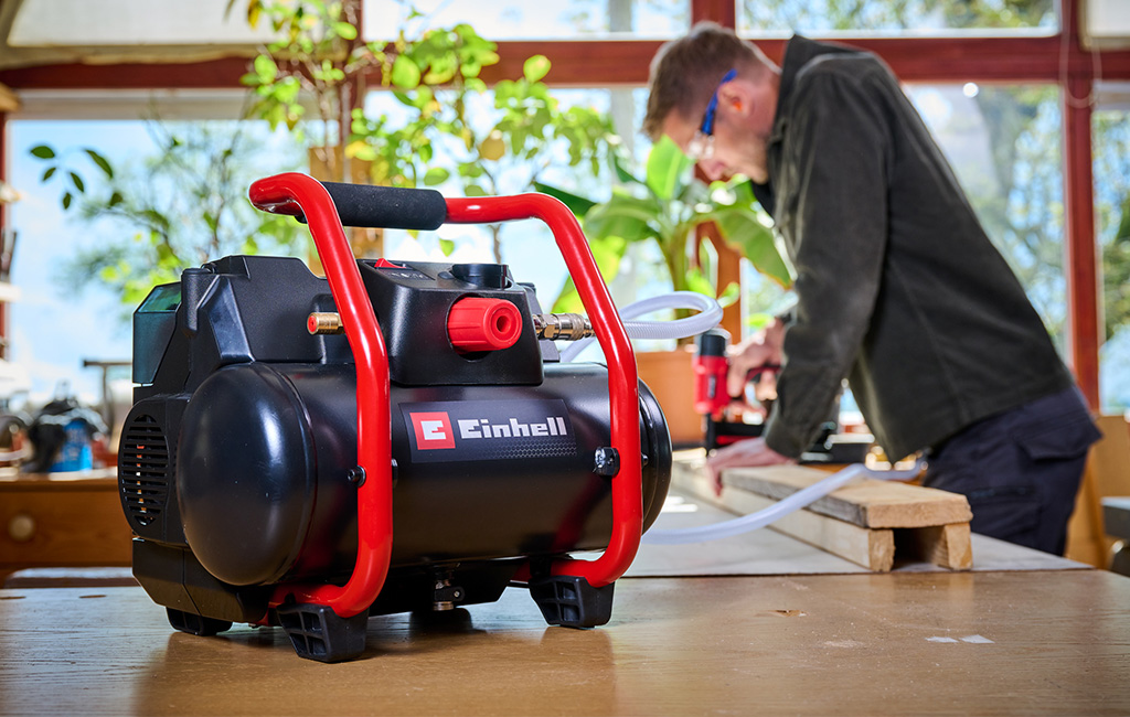 A man works in a workshop with a cordless Einhell compressor on a table in the foreground.