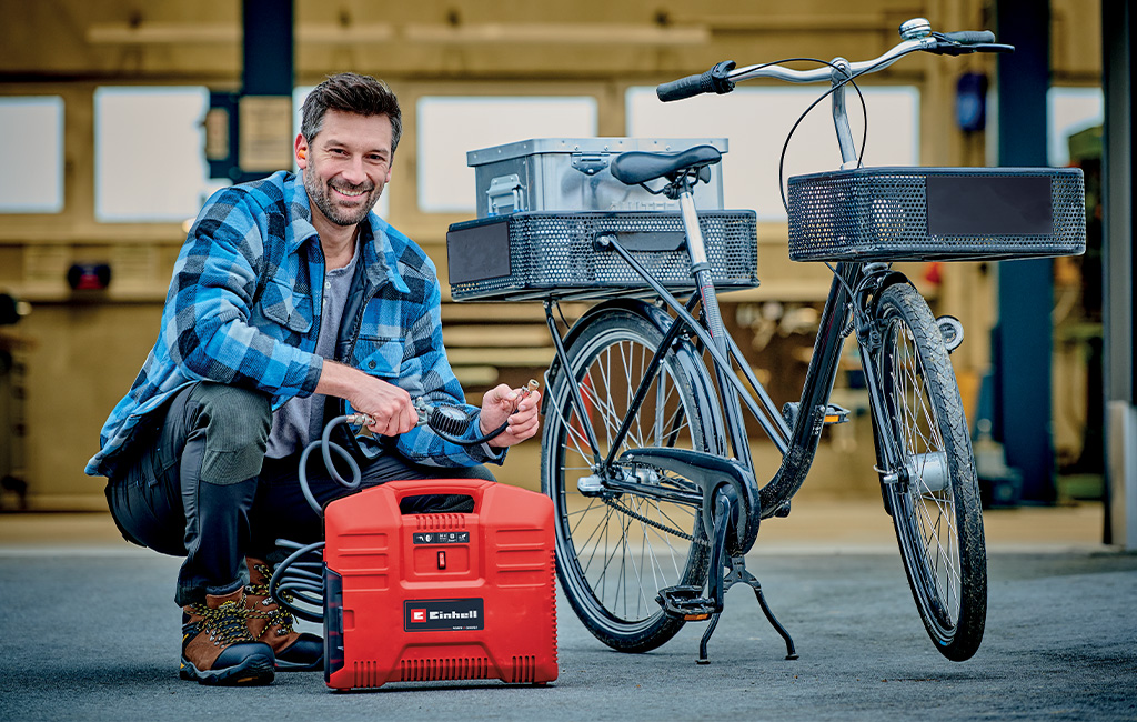 A man uses a Einhell cordless portable compressor to inflate bicycle tires in a workshop.