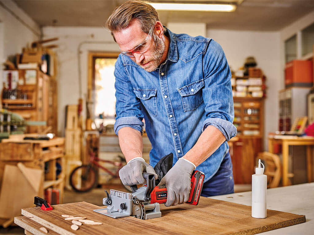 A man is using the Einhell cordless biscuit jointer on a wooden board. Biscuit dowels and wood glue are ready on the table.