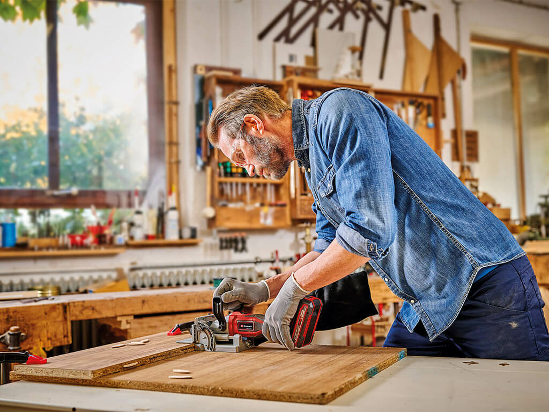 A man uses the Einhell cordless biscuit jointer in a workshop on two wooden boards. Dowels and a clamp are ready on the table.