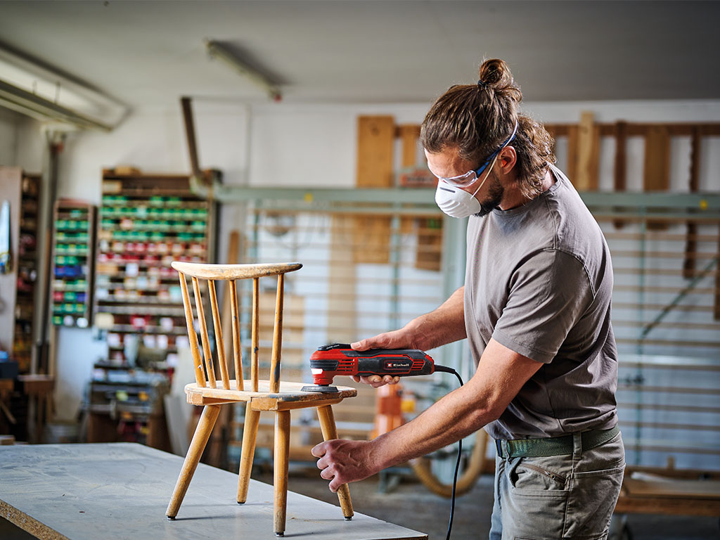 Ein Mann schleift mit dem Einhell Multifunktionstool einen Holzstuhl in einer Werkstatt.