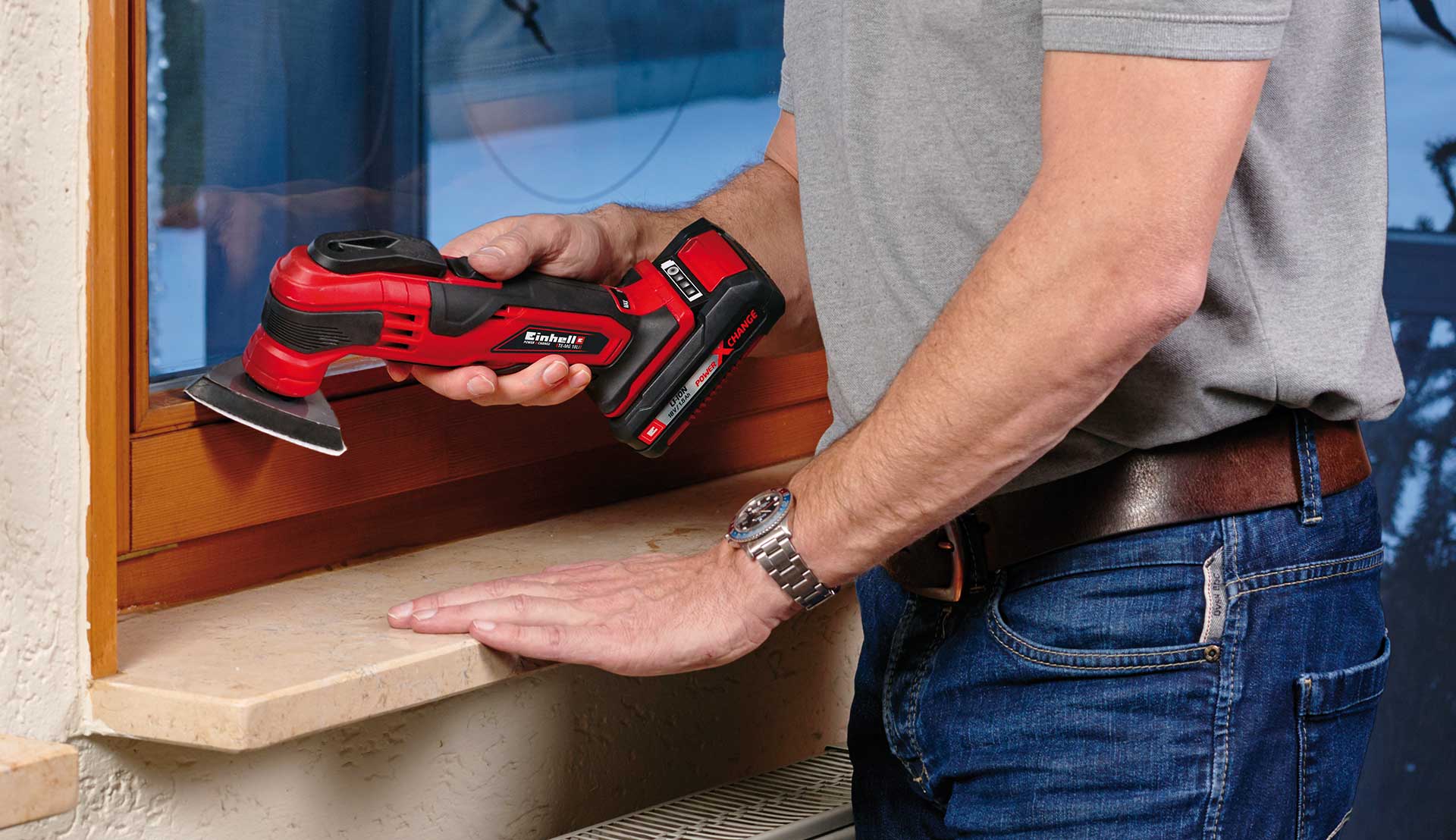 A man is using a multifunctional tool with a sanding attachment to grind a wooden window frame.