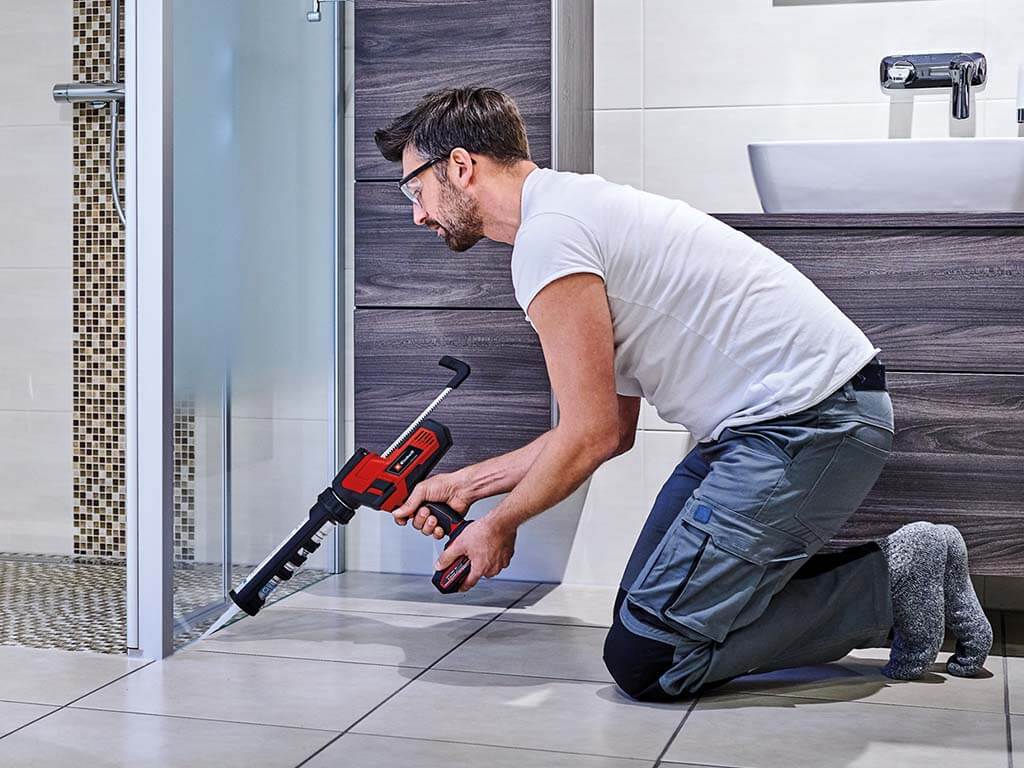 Ein Mann kniet im Bad und spritzt eine Silikonfuge zwischen Duschwand und Fliesenboden.