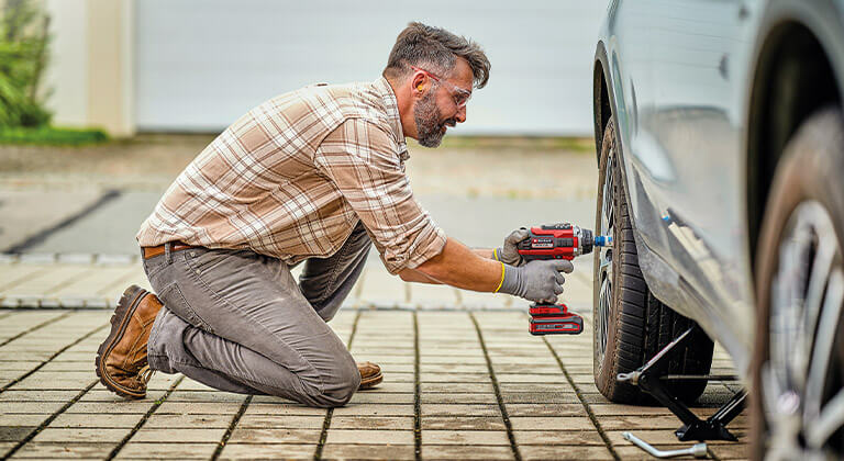 A man kneels in front of a car and loosens the wheel nuts on a tyre with an Einhell IMPAXXO cordless impact wrench.