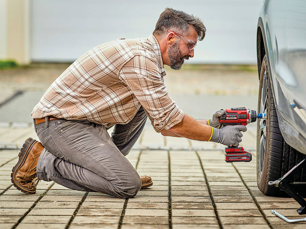 A man kneels in front of a car and loosens the wheel nuts on a tyre with an Einhell IMPAXXO cordless impact wrench.