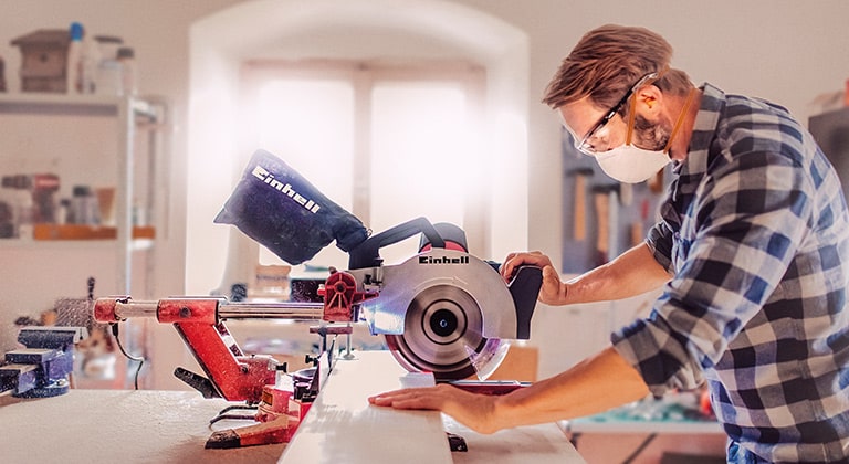 A man cuts a wooden board with an Einhell mitre saw in a workshop.