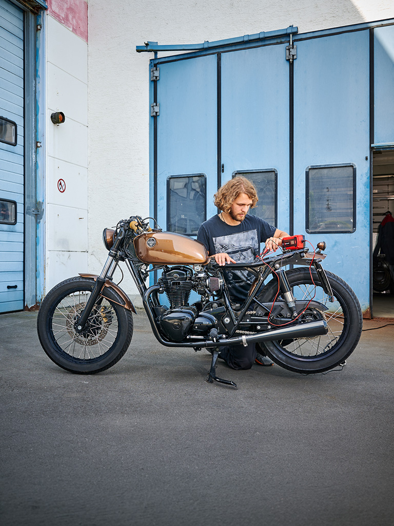 Person kneeling next to a motorcycle in front of a workshop checking the battery with an Einhell battery jump starter.