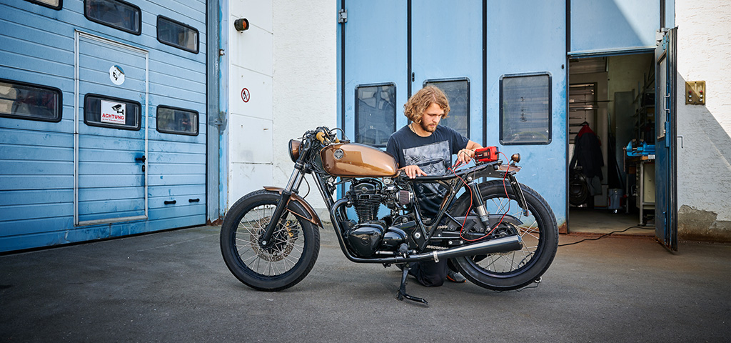 Person kneeling next to a motorcycle in front of a workshop checking the battery with an Einhell battery jump starter.