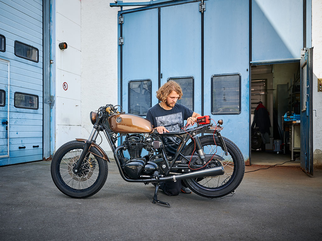 Person kneeling next to a motorcycle in front of a workshop checking the battery with an Einhell battery jump starter.