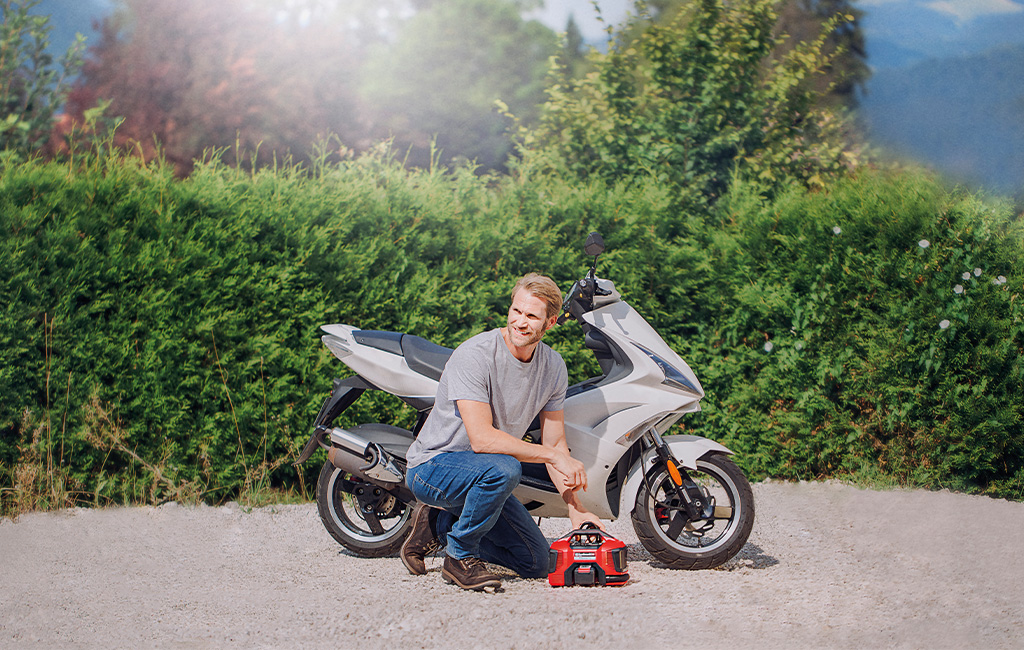 Person kneeling next to a scooter outdoors using an Einhell compressor to check the tyre pressure.