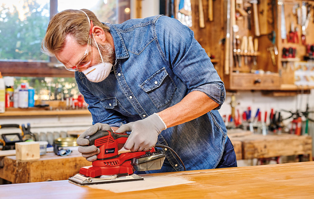Man sanding a wooden board with a rotary sander in a workshop.