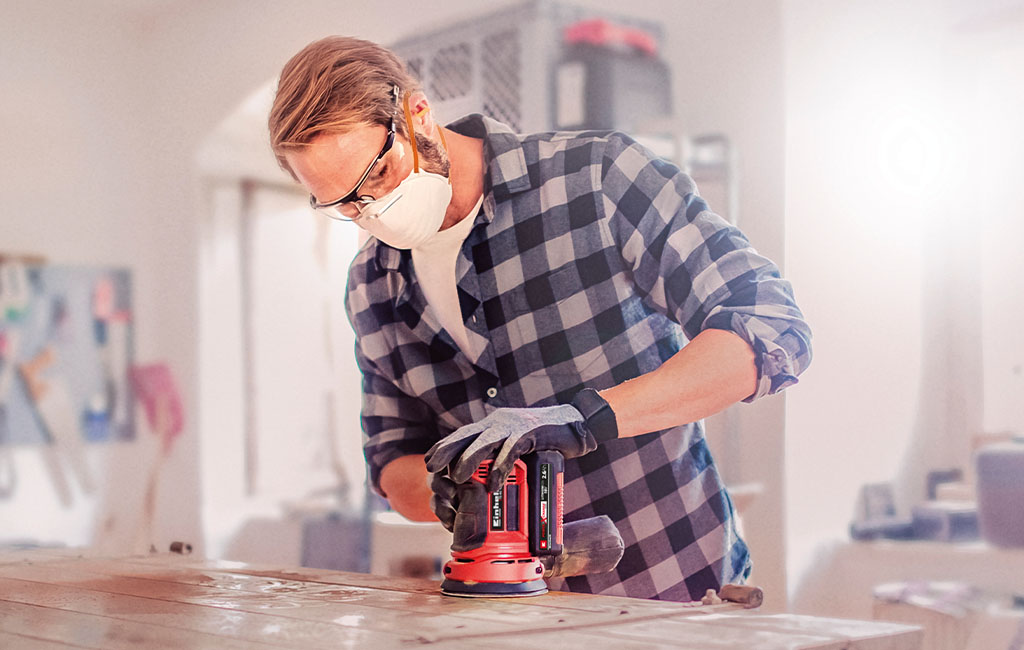 Man sanding a wooden board with a rotary sander in a workshop.