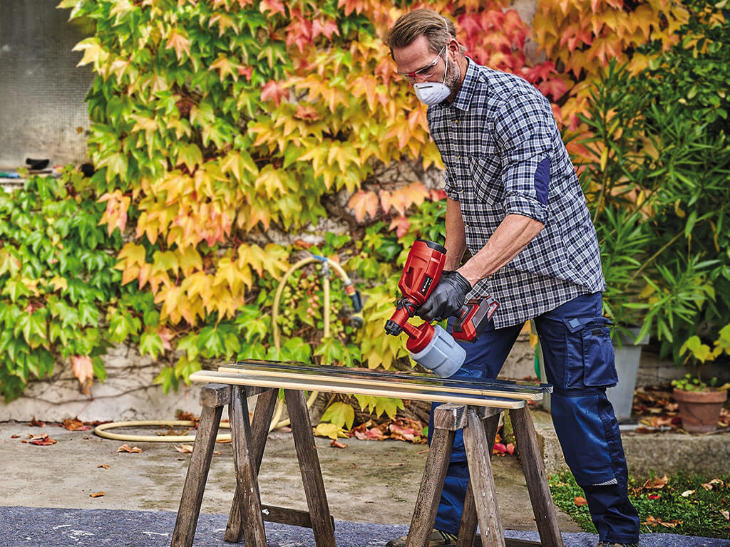 A man uses an Einhell paint sprayer to spray paint on wooden slats.