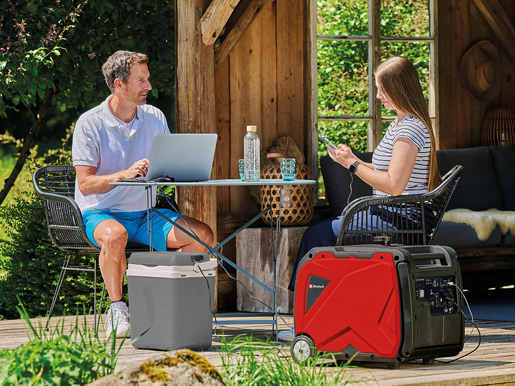 a man and a woman are sitting on a tabel in their garden working on their notebook and mobile phone connected on a power generator