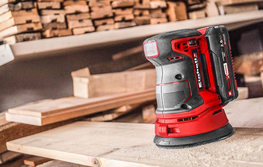 A random orbital sander sits on a wooden surface being worked on in a garage. Stacked pieces of wood are visible in the background.