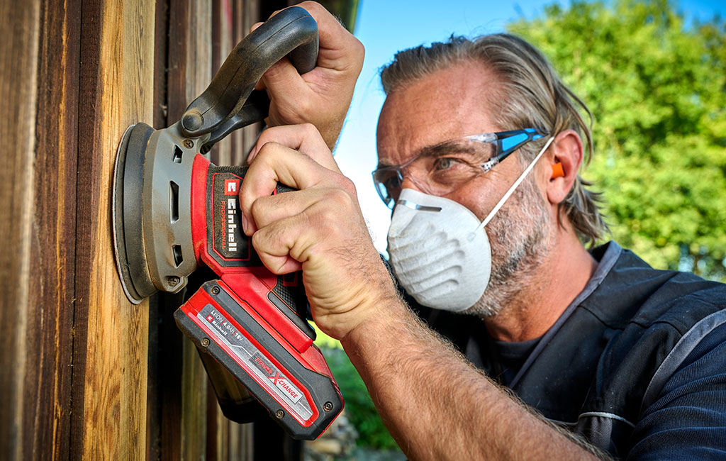 A man sands a wooden surface outdoors with a random orbital sander. He is wearing a respirator mask and hearing protection to guard against dust and noise.