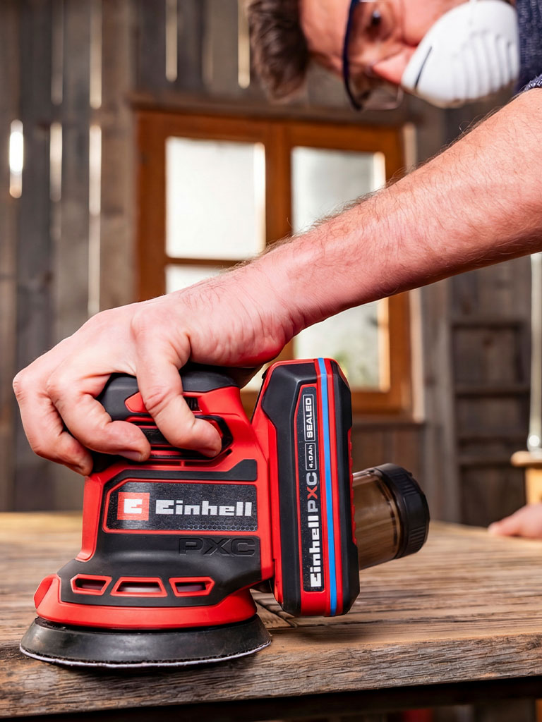 A man works on a wooden surface in a garage using a random orbital sander.