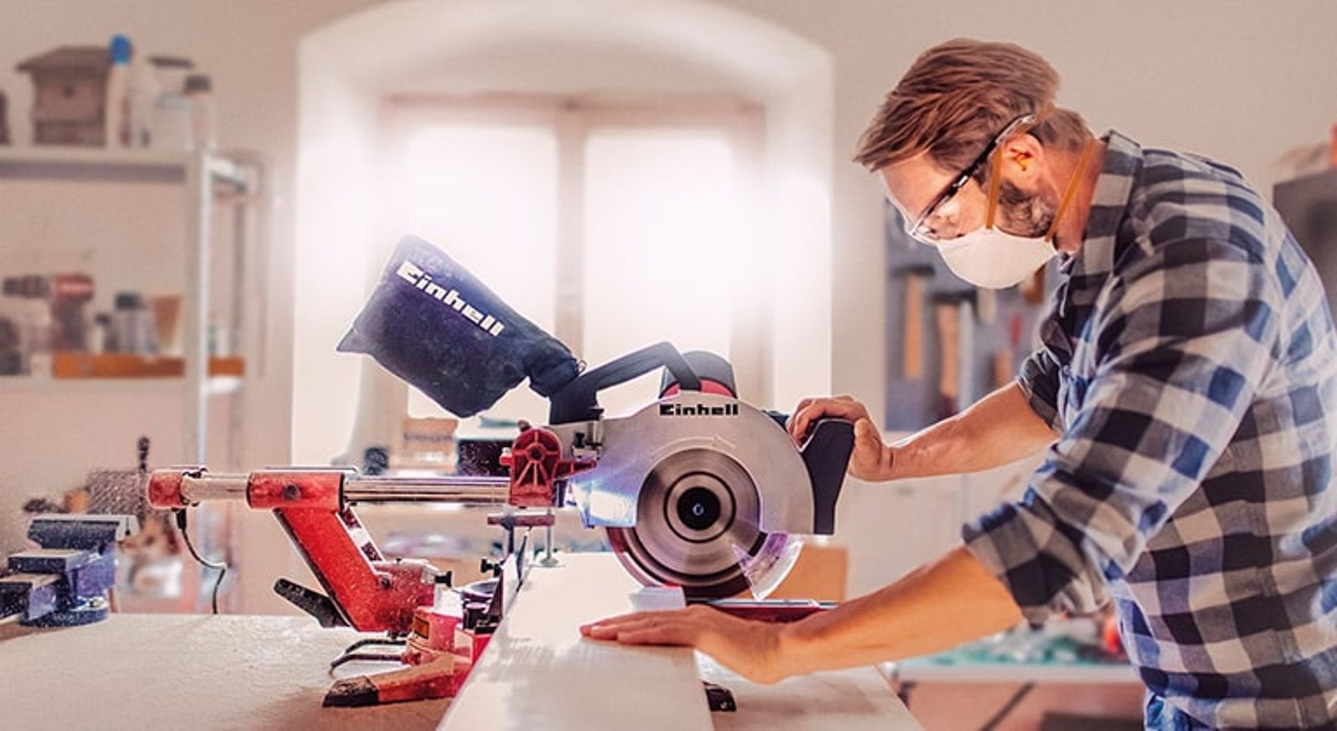 A man wearing a respiratory mask, sawing wood with a mitre saw.