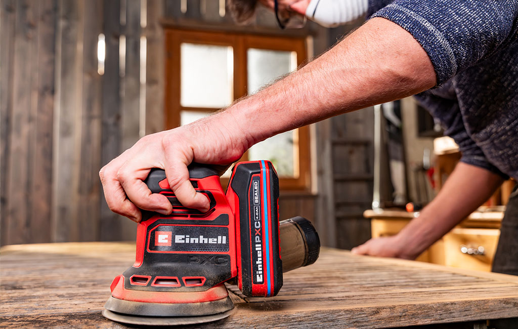An Einhell random orbital sander sands an old wooden board in a workshop.