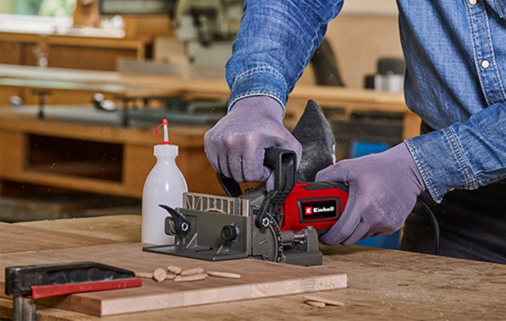 A cordless Einhell biscuit joiner cuts a slot into a wooden board while two wooden pieces are clamped together.