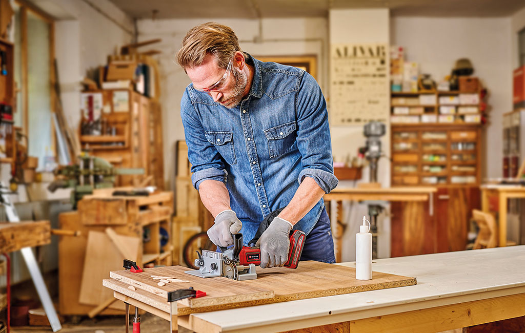 A man uses a cordless Einhell biscuit joiner to cut slots into wooden boards for a DIY project in the workshop.