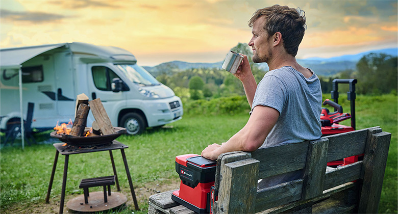Ein Mann sitzt auf einer Holzbank in der Natur, trinkt aus einem Becher und nutzt eine Einhell Akku-Kaffeemaschine. Im Hintergrund stehen ein Wohnmobil und eine Feuerstelle mit brennendem Holz.