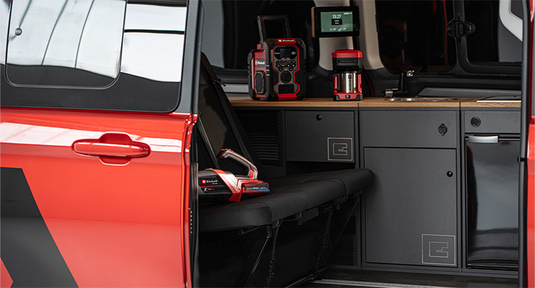 Seitlicher Blick in den Einhell-Camping-Van mit Sitzbank und Küchenzeile. Auf der Bank liegt ein Akku-Handstaubsauger mit eingesetztem Akku. Auf der Arbeitsfläche stehen ein Einhell-Akku-Radio und eine Kaffeemaschine.