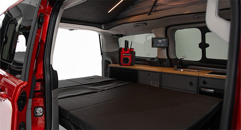 Blick in den Camping-Van mit ausgeklapptem Bett und Küchenzeile. Auf der Arbeitsfläche steht ein rotes Einhell-Akku-Radio.