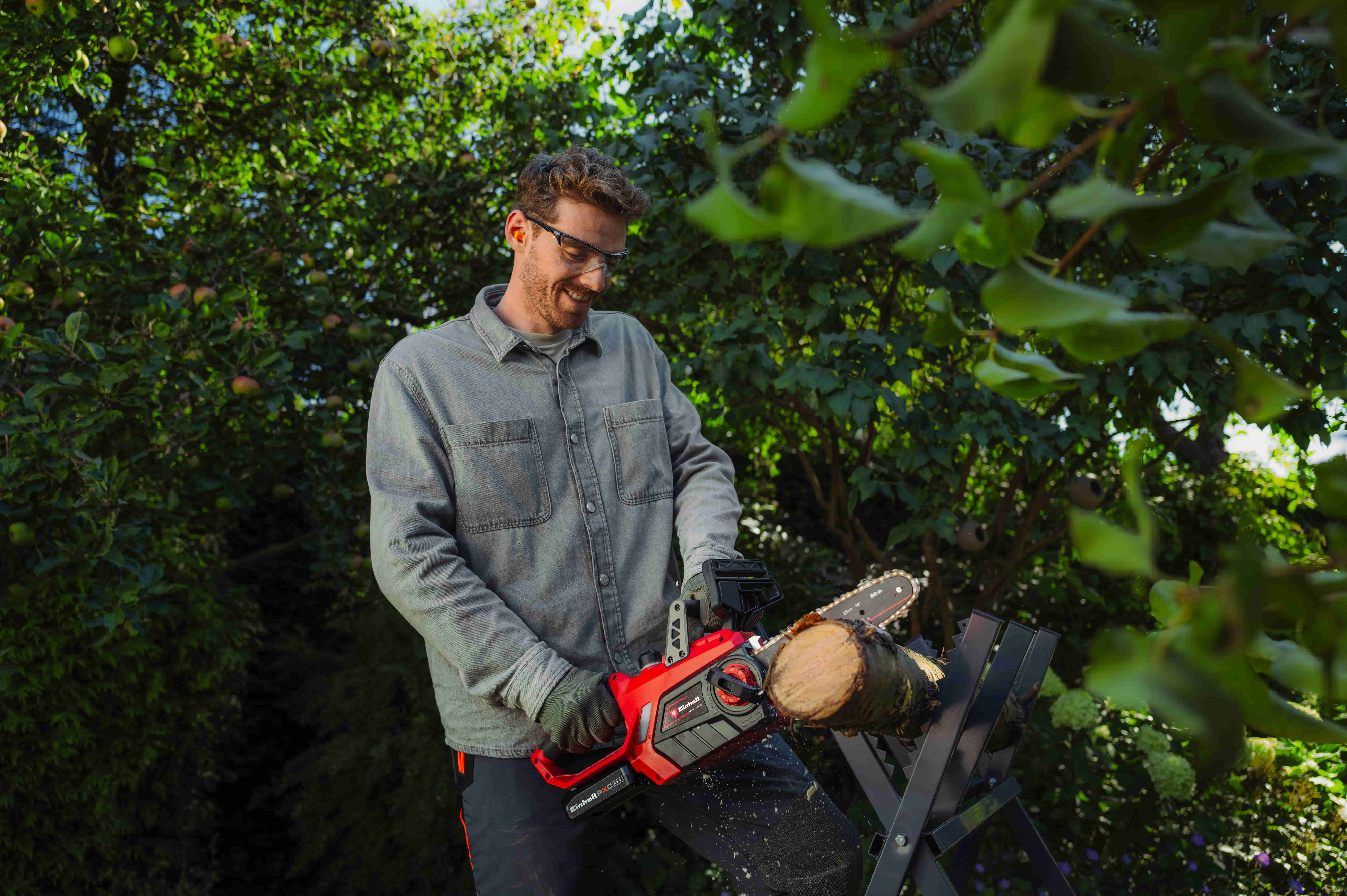 Mann mit Schutzbrille sägt im Garten einen Holzstamm mit einer roten Einhell Akku-Kettensäge auf einem Sägebock.