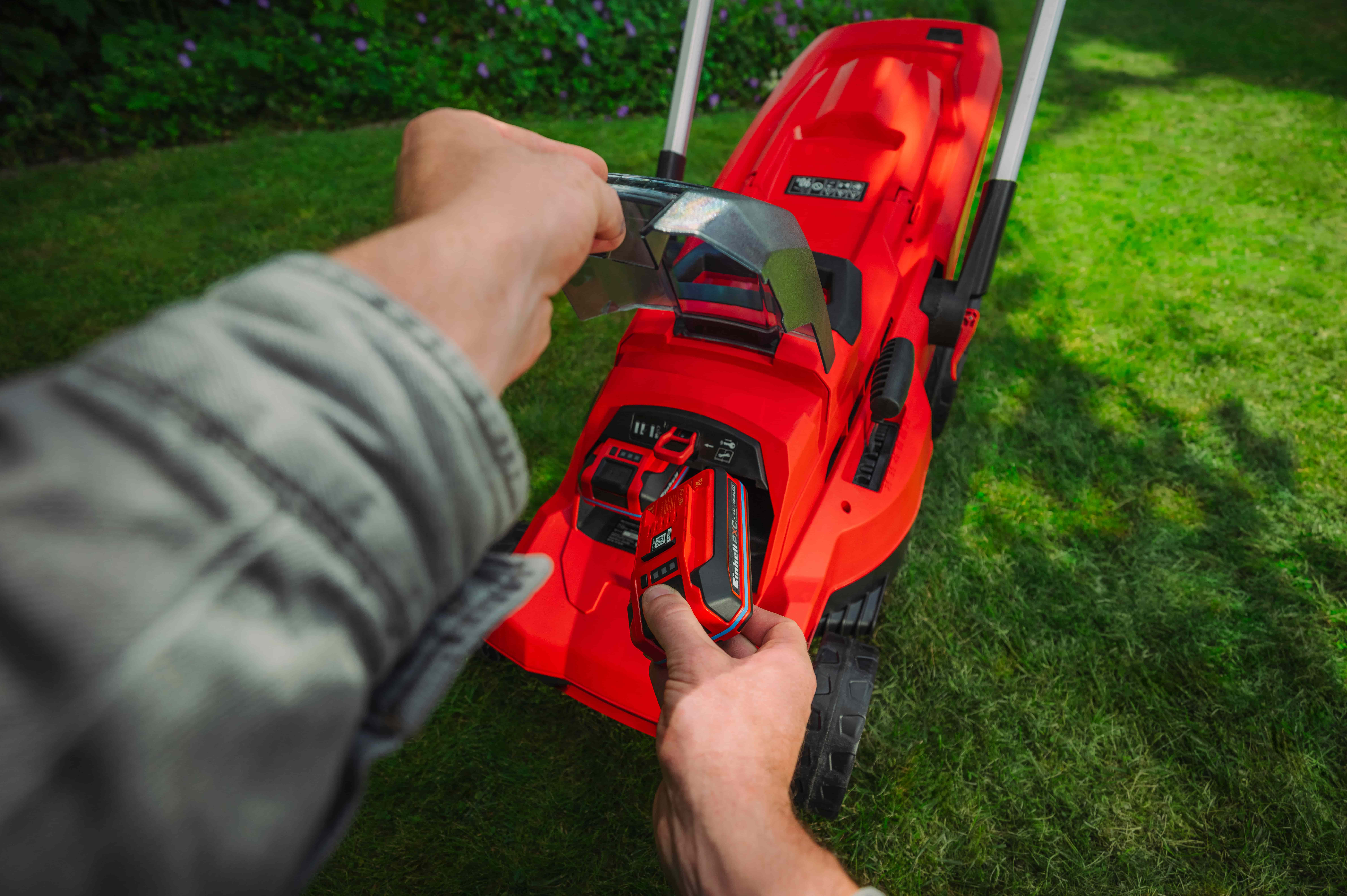 Eine Hand setzt einen Einhell Power X-Change Akku in einen roten Einhell Akku-Rasenmäher auf einer Rasenfläche ein.