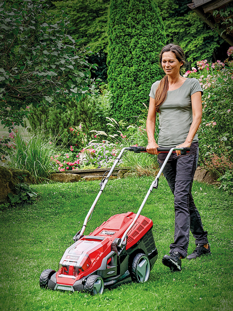 Eine Frau mäht mit einem roten Einhell-Rasenmäher den Rasen in einem grünen, blühenden Garten.