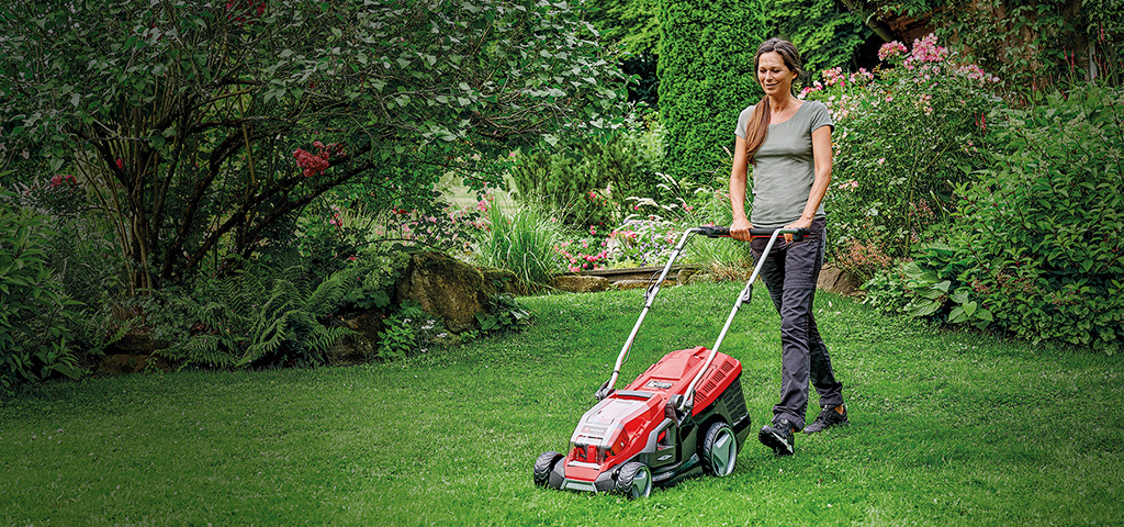 Eine Frau mäht mit einem roten Einhell-Rasenmäher den Rasen in einem grünen, blühenden Garten.