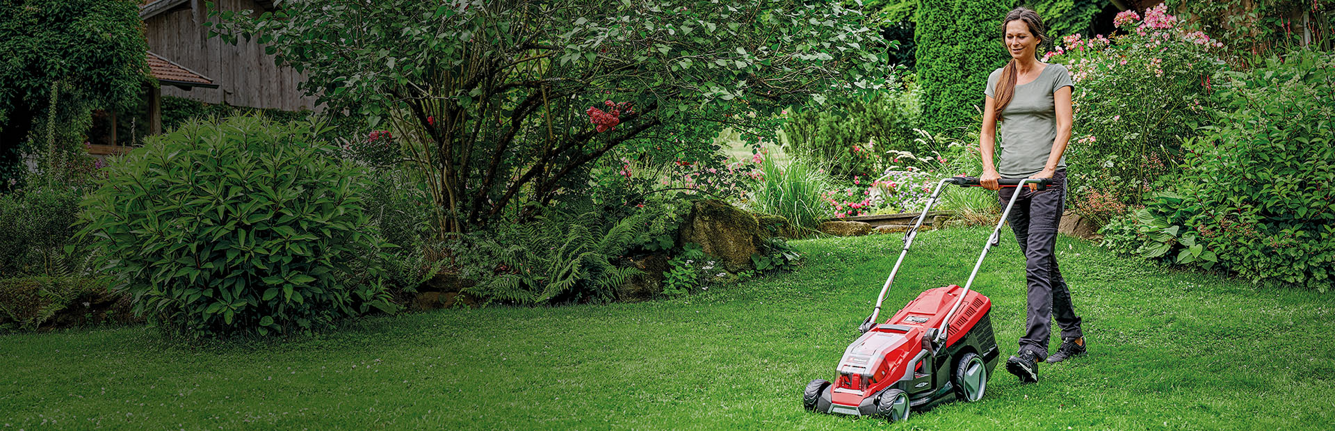 Eine Frau mäht mit einem roten Einhell-Rasenmäher den Rasen in einem grünen, blühenden Garten.