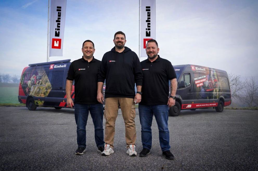 Three men in black hoodies stand in front of two Einhell vans with Einhell flags in the background.