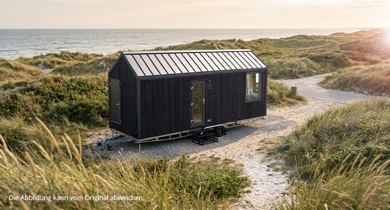 Ein schwarzes Tiny House auf einem Anhänger, geparkt auf einem sandigen Weg inmitten einer Dünenlandschaft am Meer.