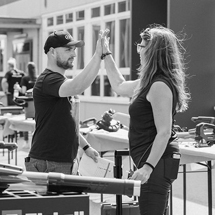 Ein Mann und eine Frau geben sich ein High-Five bei einer Einhell-Veranstaltung im Freien mit Werkzeugaustellung, in Schwarz-Weiß dargestellt.