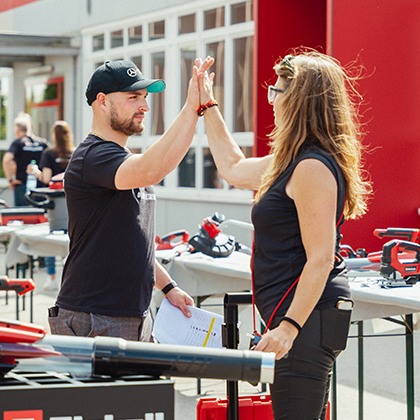 Ein Mann und eine Frau geben sich ein High-Five bei einer Einhell-Veranstaltung im Freien mit Werkzeugaustellung.