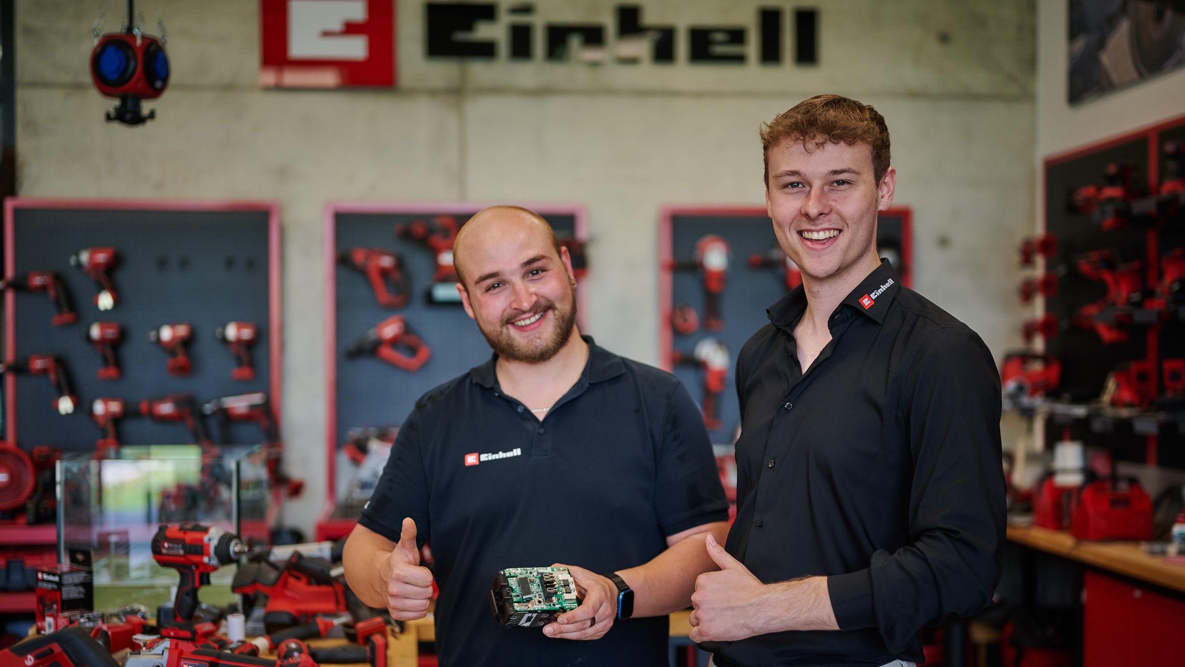 Two men being surrounded by Einhell products and smiling into the camera.