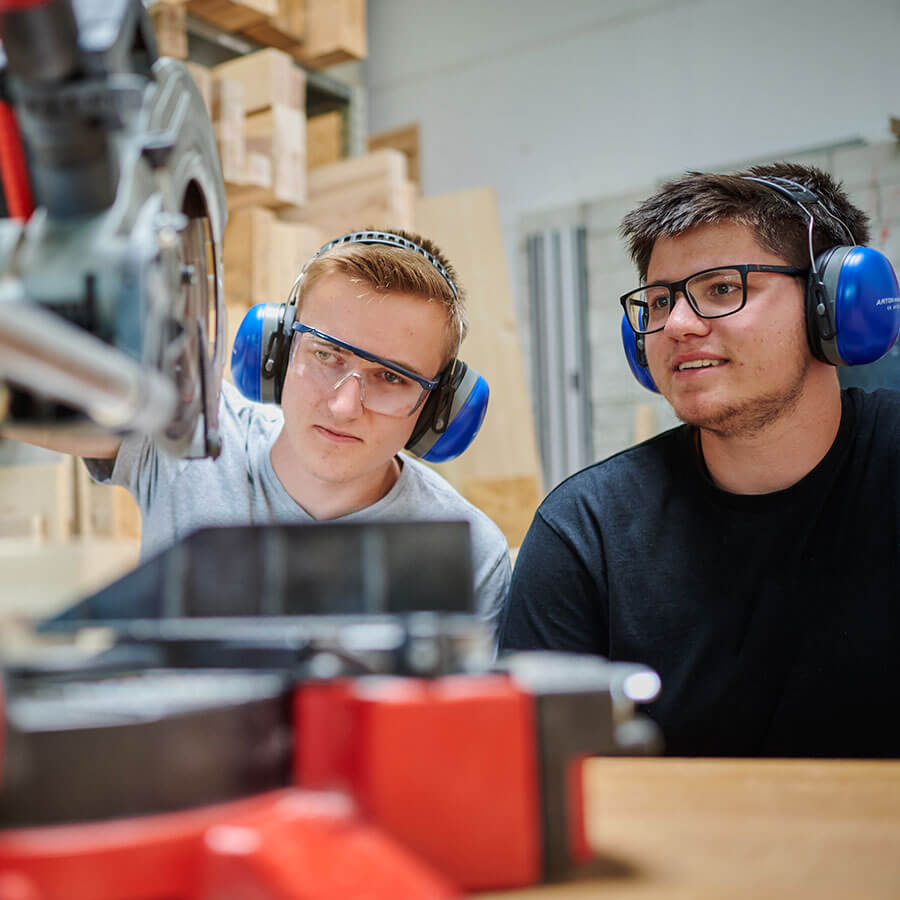 two men with earmuffs on cutting something with a table saw