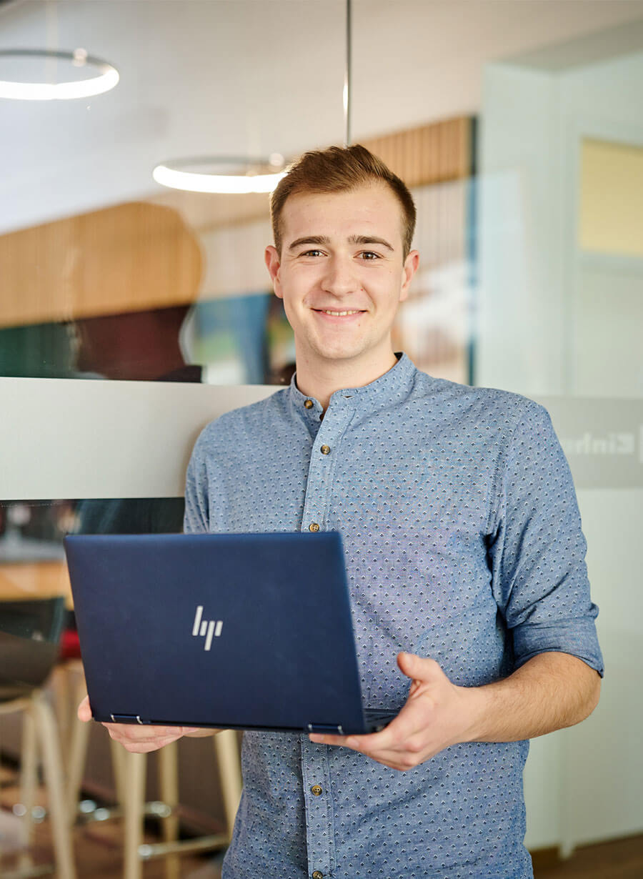 A man is holding a laptop and smiles into the camera.