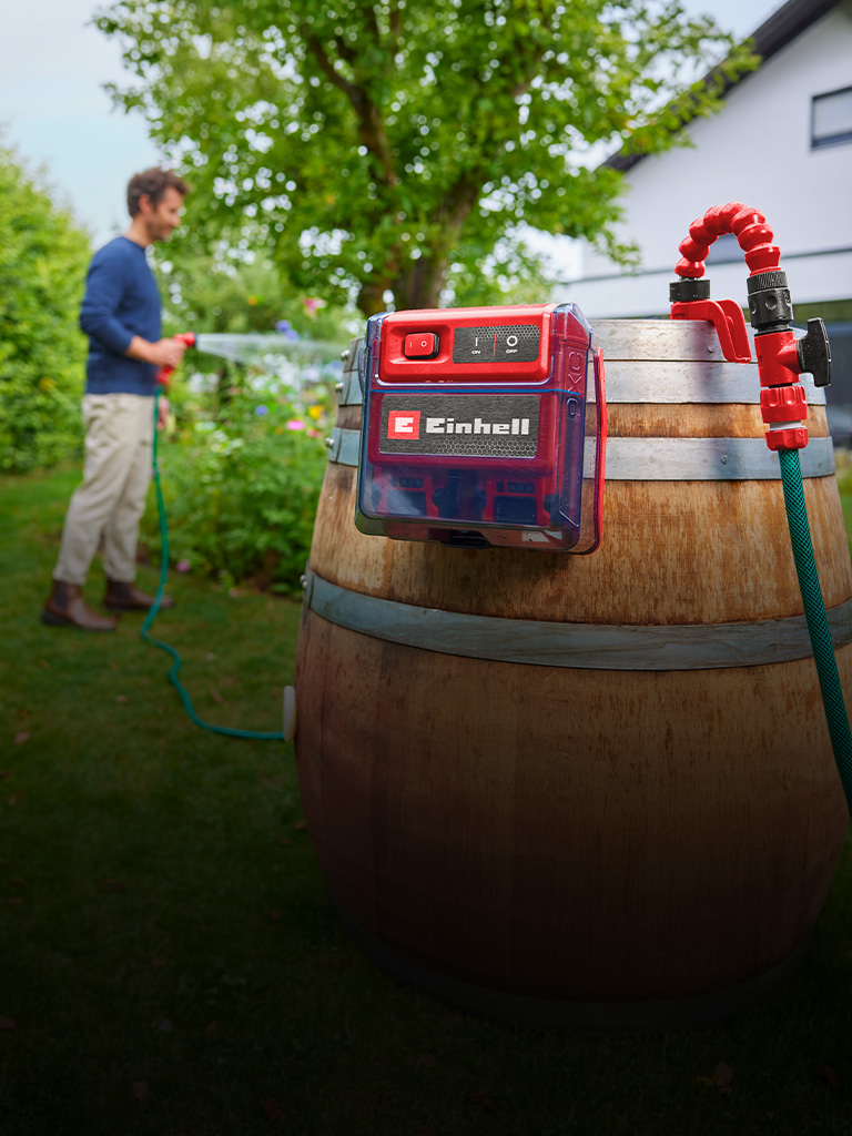 Einhell Akku-Klarwasserpumpe an einem Holzfass im Garten, während eine Person im Hintergrund Pflanzen bewässert.