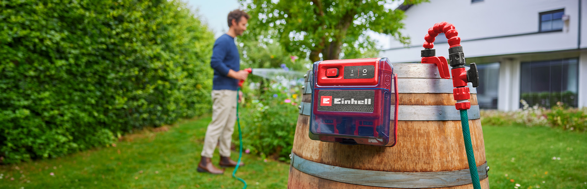 Einhell Akku-Klarwasserpumpe an einem Holzfass im Garten, während eine Person im Hintergrund Pflanzen bewässert.