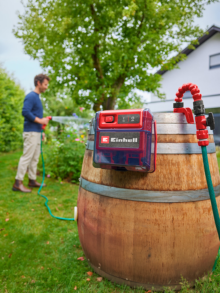 Einhell Akku-Klarwasserpumpe an einem Holzfass im Garten, während eine Person im Hintergrund Pflanzen bewässert.