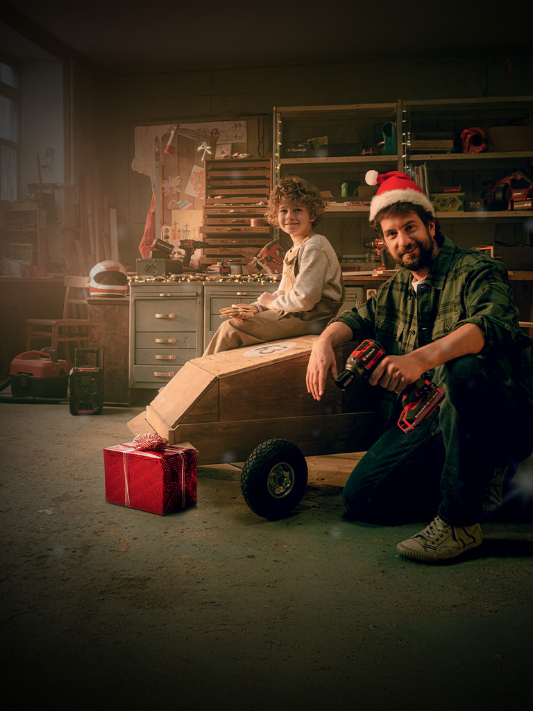 Ein Kind sitzt auf einem Holzauto, ein Mann mit Weihnachtsmütze kniet daneben mit einem Akkuschrauber. Im Hintergrund ist eine Werkbank, daneben ein Geschenk in rotem Papier.