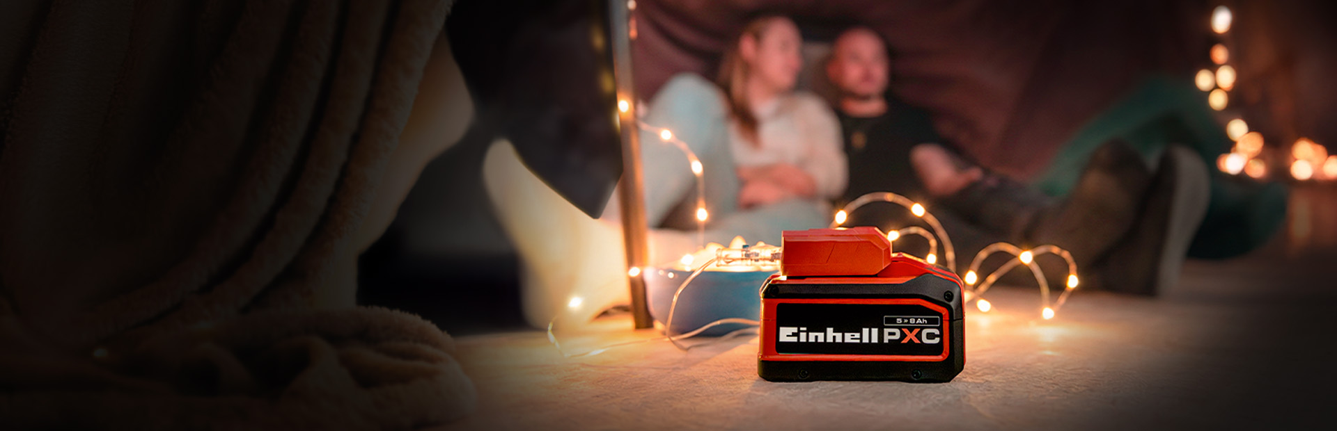 An Einhell Power X-Change battery (5.2 Ah) in the foreground, with a couple sitting in a cozy setting with fairy lights in the background.