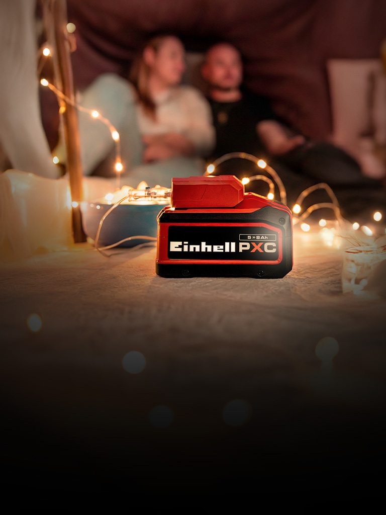 An Einhell Power X-Change battery (5.2 Ah) in the foreground, with a couple sitting in a cozy setting with fairy lights in the background.