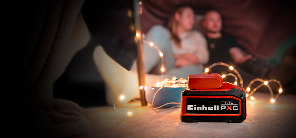 An Einhell Power X-Change battery (5.2 Ah) in the foreground, with a couple sitting in a cozy setting with fairy lights in the background.