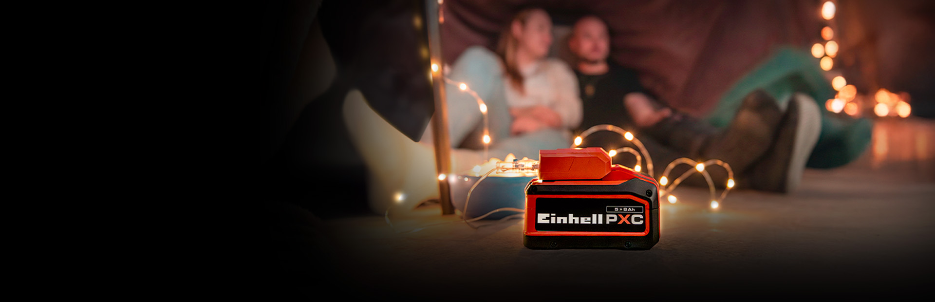 An Einhell Power X-Change battery (5.2 Ah) in the foreground, with a couple sitting in a cozy setting with fairy lights in the background.