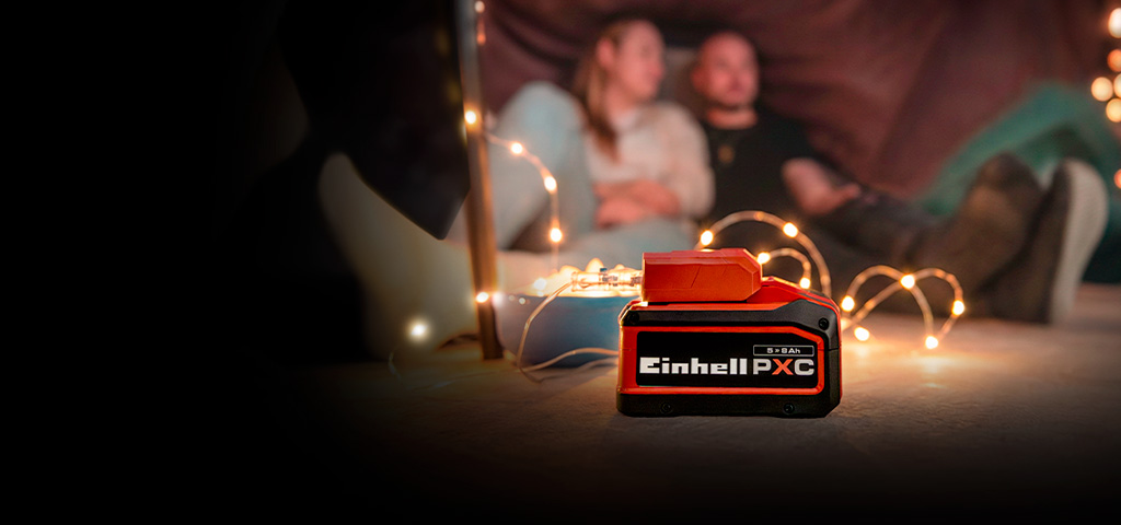 An Einhell Power X-Change battery (5.2 Ah) in the foreground, with a couple sitting in a cozy setting with fairy lights in the background.